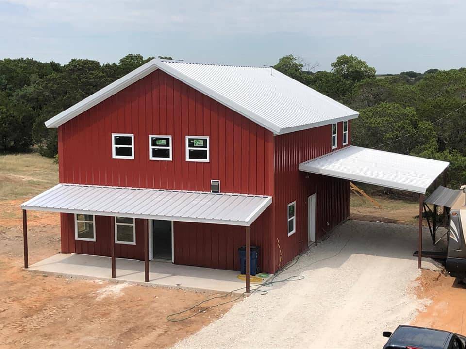 Red two-story steel barndominium with wraparound porch and attached carport in Texas